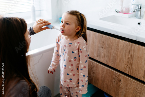 4-year-old girl in pajamas stands in bathroom while mother carefully brushes her teeth with electric toothbrush. Everyday routine reflects parent-child connection, healthy habits, and nurturing care.
 photo