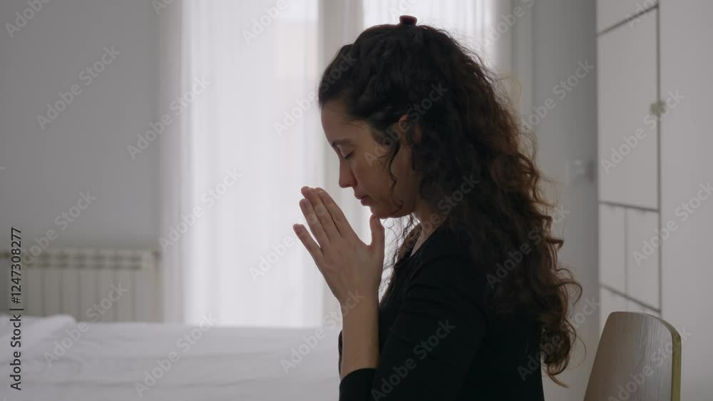 Profile shot of young woman praying in bedroom with bright daylight in room. Her eyes closed and she speaks quietly to herself. Worship and faith practice in house or apartment dreamy look Profile shot of young woman praying in bedroom with bright daylight in room. Her eyes closed and she speaks quietly to herself. Worship and faith practice in house or apartment dreamy look