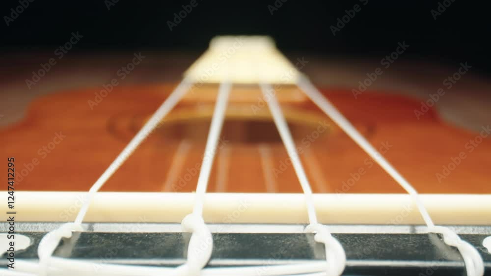 Zoom in on wooden ukulele with white nylon strings on dark background. Close-up macro shot of musical string instrument with fretboard on a brown table. Concept of learning to play hawaiian guitar Zoom in on wooden ukulele with white nylon strings on dark background. Close-up macro shot of musical string instrument with fretboard on a brown table. Concept of learning to play hawaiian guitar