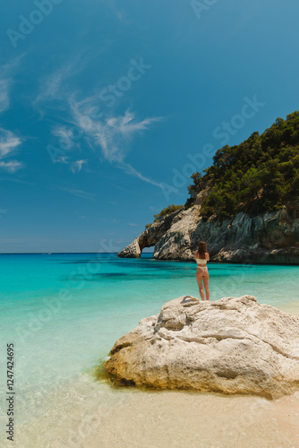 Fototapeta Naklejka Na Ścianę i Meble -  Woman standing on a rock admiring the stunning turquoise water and natural arch of cala goloritze beach in sardinia, italy