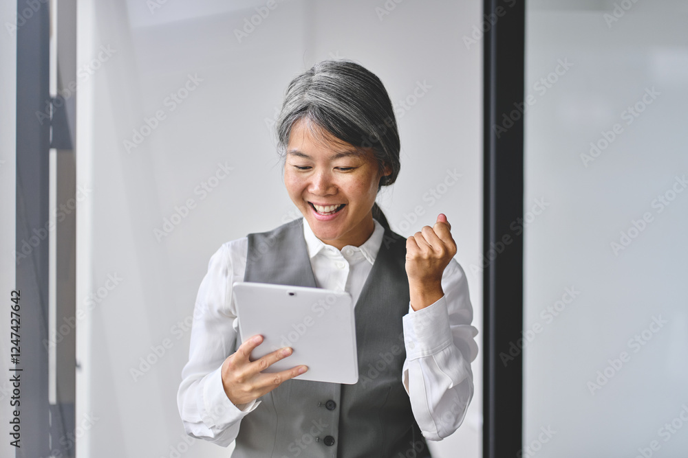 Happy excited professional older mature Asian business woman employee, executive manager looking at digital tablet feeling joy celebrating profit, corporate success standing at work in office. Happy excited professional older mature Asian business woman employee, executive manager looking at digital tablet feeling joy celebrating profit, corporate success standing at work in office.
