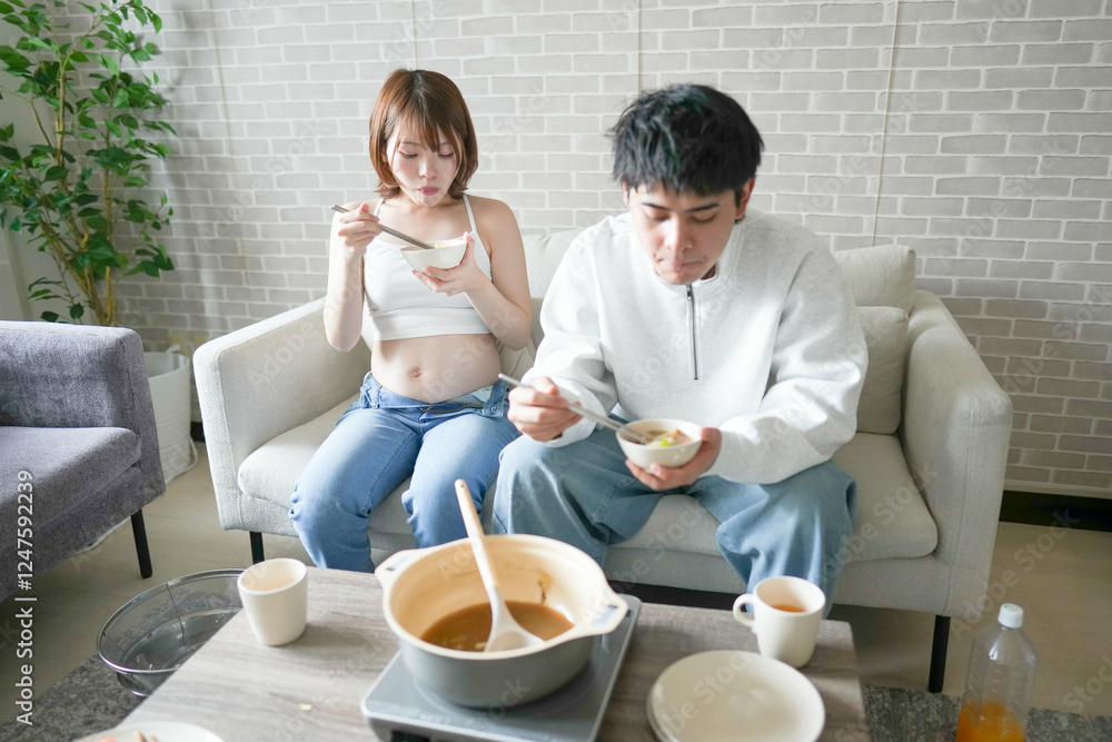 A Japanese pregnant woman in her 20s, wearing a dress that shows her stomach, sits on a sofa in an apartment and her husband eats a hot pot dish. A Japanese pregnant woman in her 20s, wearing a dress that shows her stomach, sits on a sofa in an apartment and her husband eats a hot pot dish.