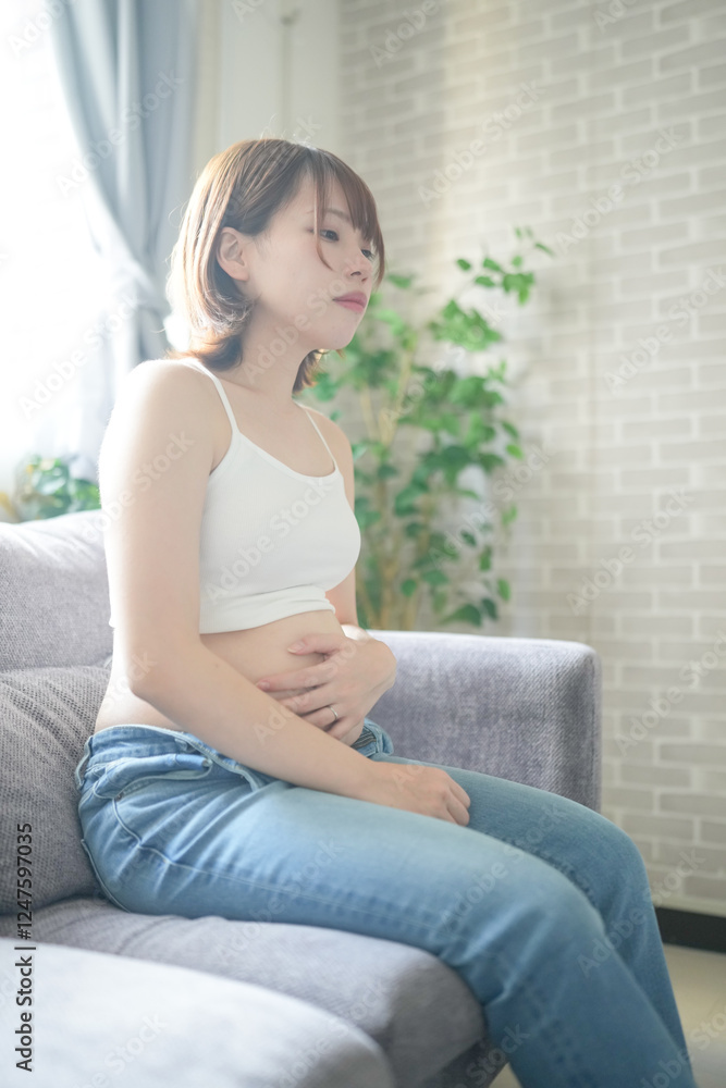 A pregnant Japanese woman in her 20s wearing clothes that show her stomach is sitting on a sofa in her apartment, holding her big belly. A pregnant Japanese woman in her 20s wearing clothes that show her stomach is sitting on a sofa in her apartment, holding her big belly.