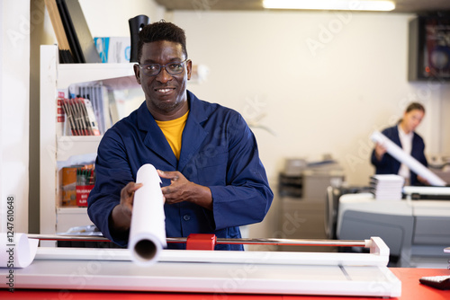 Excited middle-aged African American male typographer in uniform showing large format paper in a roll in the typography photo