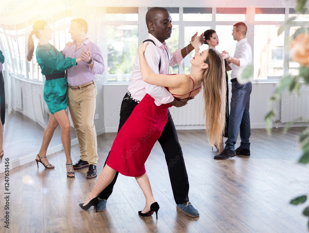 Fototapeta premium Woman in elegant evening dress dances incendiary tango with African American male partner during choreography lesson. Group training and rehearsal, preparation for competitions