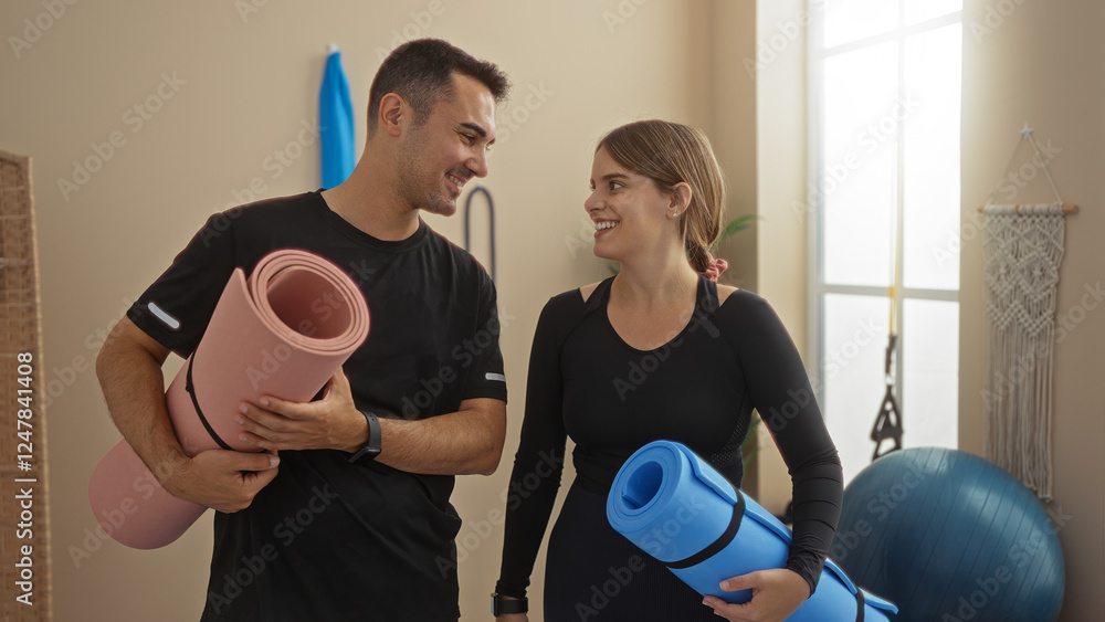 Man and woman smiling together in gym holding yoga mats suggesting a healthy, active lifestyle in an indoor sports center setting. Man and woman smiling together in gym holding yoga mats suggesting a healthy, active lifestyle in an indoor sports center setting.