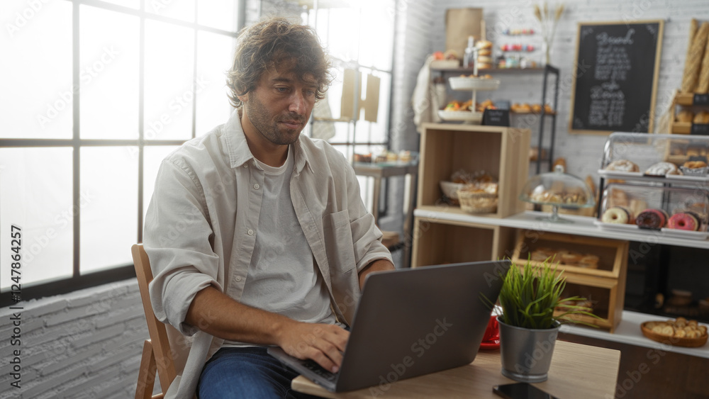 Young man working on laptop in a cozy bakery surrounded by delicious pastries and bread. Young man working on laptop in a cozy bakery surrounded by delicious pastries and bread.
