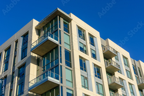 Exterior view of upscale modern luxury apartment building with floor to ceiling windows and glass railed balconies against vibrant blue sky. Looking up.