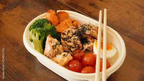 A healthy bento box with grilled salmon, steamed broccoli, cherry tomatoes, roasted sweet potatoes, and a boiled egg, served with chopsticks on a rustic wooden table.