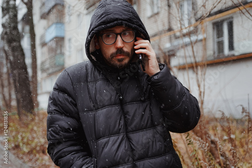 A young man in casual style walks through the snowy city on a winter day, chatting on his phone and sipping coffee. The lifestyle photo captures his relaxed, modern vibe and cheerful energy in an urba photo