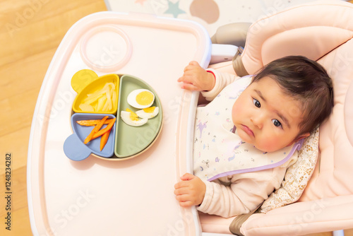 Precious six-month-old Japanese Asian baby sitting in her highchair to eat a nutritious meal of fruit pieces, vegetables and boiled egg on a colorful tricolor plate. BLW method