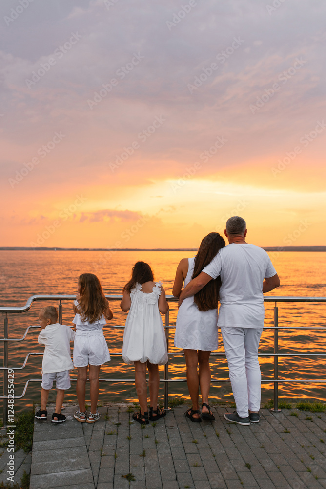 Fototapeta premium Happy family of five is standing on pier overlooking the ocean and enjoying the view and taking in the peaceful atmosphere