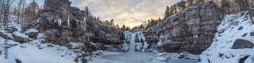 Frozen waterfall surrounded by snow-covered rocky cliffs and bare trees at sunset. Concept of winter wonderland. For landscape photography.