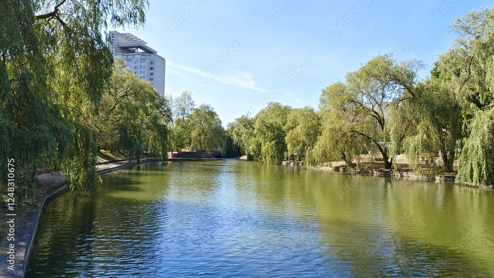 Willows and other deciduous trees grow on the grassy concreted banks of the canal, and buildings stand. A pedestrian walkway with a stone parapet runs nearby. Sunny autumn weather and blue sky Willows and other deciduous trees grow on the grassy concreted banks of the canal, and buildings stand. A pedestrian walkway with a stone parapet runs nearby. Sunny autumn weather and blue sky