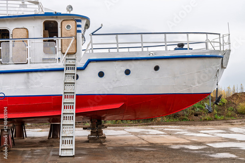 White and red ship raised on blocks for maintenance at dry dock near river, with cloudy sky and autumn trees in background. Concept of ship repair