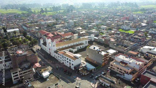 Aerial orbit shot of Machachi city, focusing on its main church and central park, surrounded by urban and rural landscapes. Located in Pichincha province, Ecuador