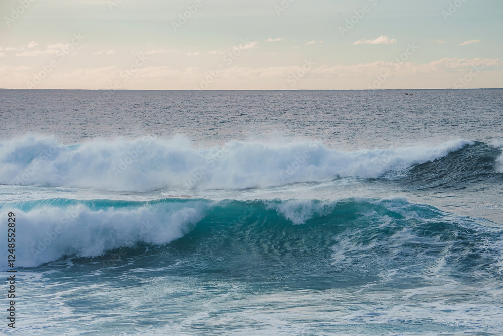 Large turquoise waves roll in the foreground, a small boat is visible on the horizon, and pastel toned clouds fill the sky in a coastal Hawaii scene. Large turquoise waves roll in the foreground, a small boat is visible on the horizon, and pastel toned clouds fill the sky in a coastal Hawaii scene.