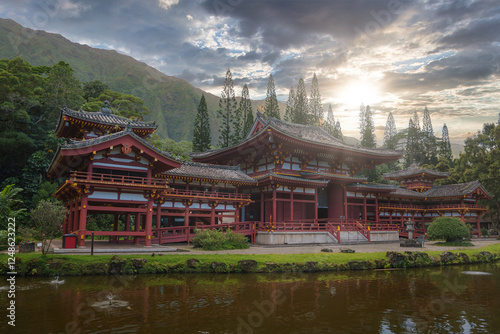 The Byodo In Temple in Oahu, Hawaii, surrounded by lush greenery, a serene pond, and dramatic mountains under a partly cloudy sky.
