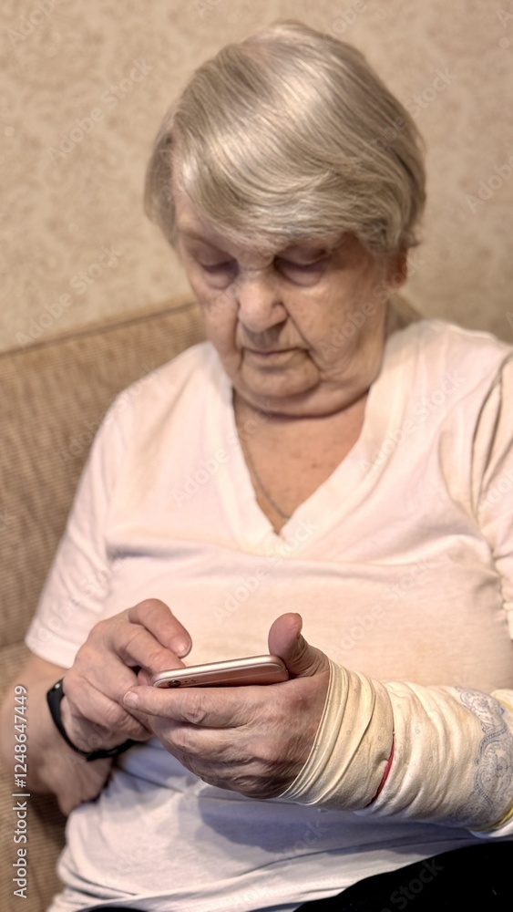 Elderly woman with broken arm width cast for the arm sitting at beige couch holding smartphone in her hand looking for important information using rose smartphone Elderly woman with broken arm width cast for the arm sitting at beige couch holding smartphone in her hand looking for important information using rose smartphone