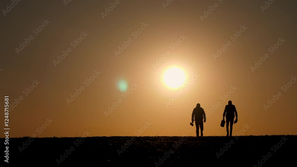 Silhouettes of two farmers walking across a field during sunrise or sunset, carrying tools and a bucket. Silhouettes of two farmers walking across a field during sunrise or sunset, carrying tools and a bucket.