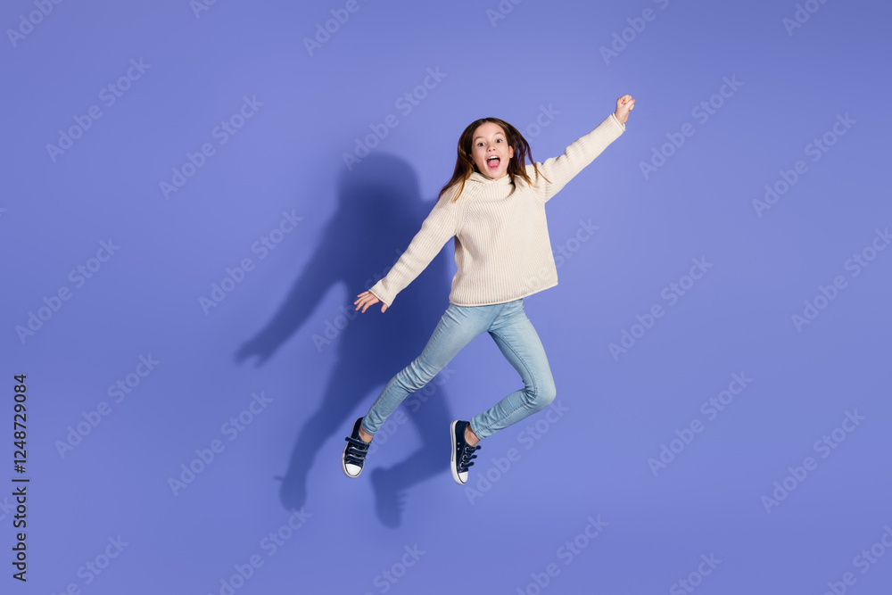Obraz premium Playful young girl jumping against a purple background in casual sweater and jeans showcasing energetic fun