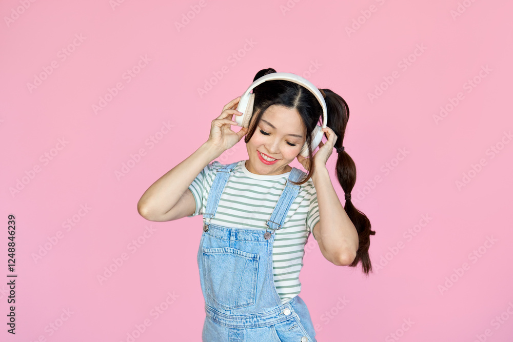 Happy funky young Asian woman, cool girl wearing striped t-shirt denim dress and headphones enjoying listening trendy pop music dancing having fun isolated on pink background. Happy funky young Asian woman, cool girl wearing striped t-shirt denim dress and headphones enjoying listening trendy pop music dancing having fun isolated on pink background.