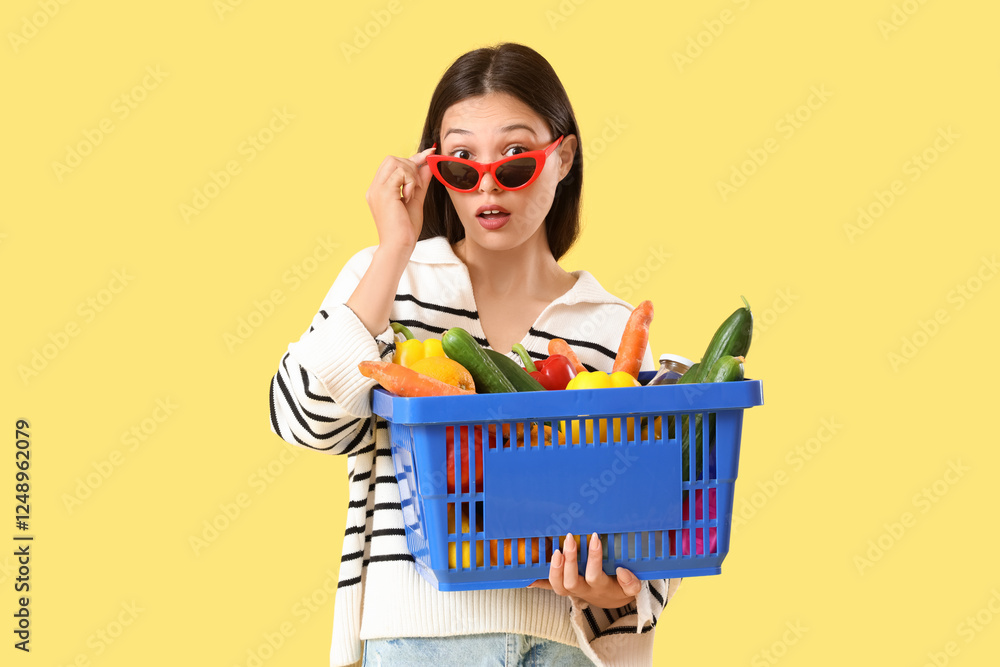 Shocked young Asian woman in sunglasses with full shopping basket on yellow background Shocked young Asian woman in sunglasses with full shopping basket on yellow background