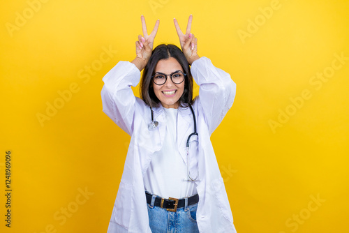 Young brunette doctor woman wearing stethoscope standing over isolated yellow background Posing funny and crazy with fingers on head as bunny ears, smiling cheerful