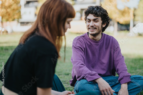 A young couple sitting on grass in a park, talking and enjoying a sunny day. Their expressions show happiness and ease, creating a warm and friendly atmosphere.