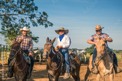 Portrait of 3 young cowboys riding their horses under a blue sky and surrounded by green vegetation. Front view.