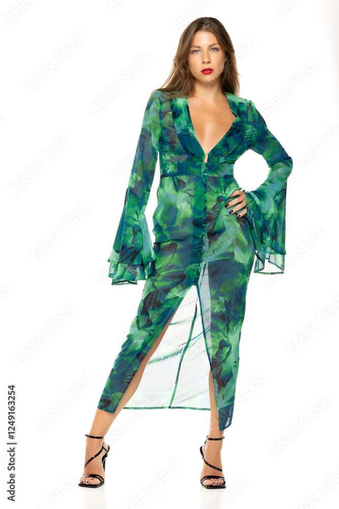 Young woman in green and blue floral maxi dress with plunging neckline and bell sleeves. Hands on hips, confident pose. Full-body shot against white background. Wearing black sandals Young woman in green and blue floral maxi dress with plunging neckline and bell sleeves. Hands on hips, confident pose. Full-body shot against white background. Wearing black sandals