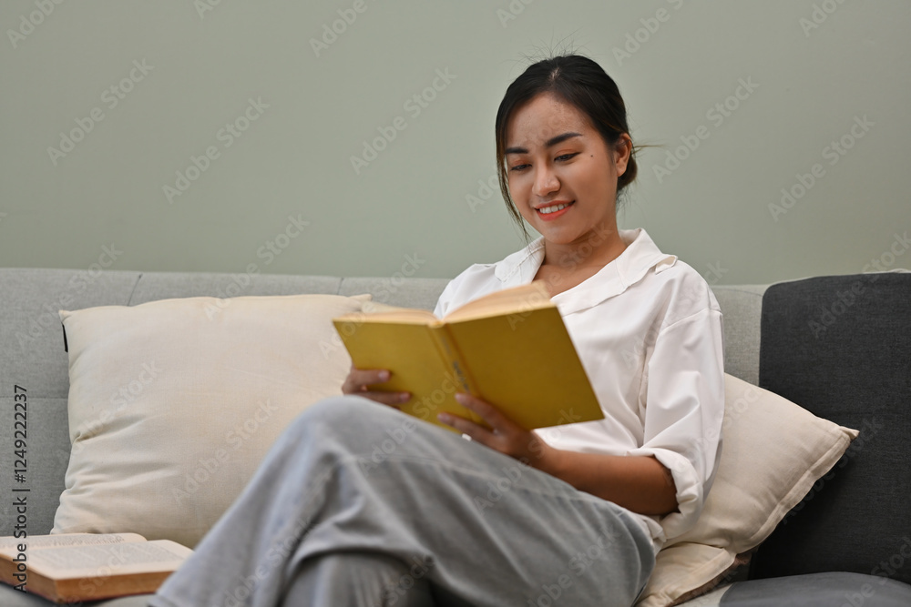 A young adult woman sits comfortably on a couch, smiling as she reads a book, surrounded by cozy pillows and a calming atmosphere, representing relaxation, learning, and mindfulness A young adult woman sits comfortably on a couch, smiling as she reads a book, surrounded by cozy pillows and a calming atmosphere, representing relaxation, learning, and mindfulness