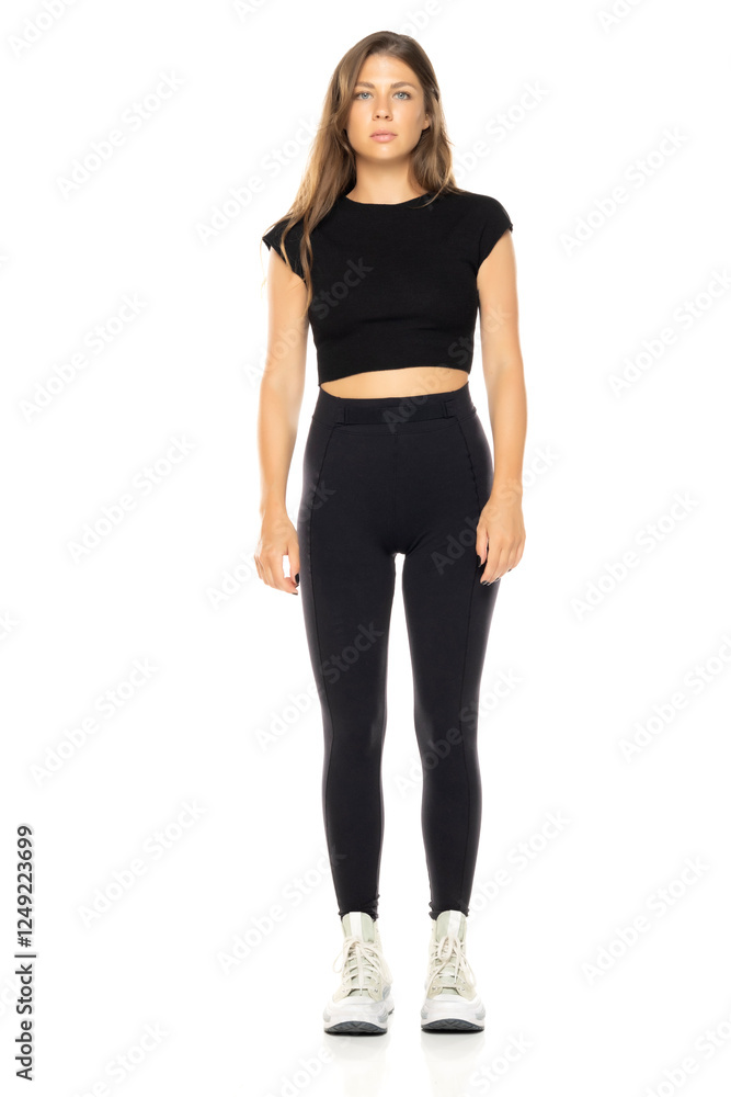 Fototapeta premium Young woman in black crop top and high-waisted leggings. Long brown hair. Full-body shot against white background. Wearing white sneakers. Neutral expression