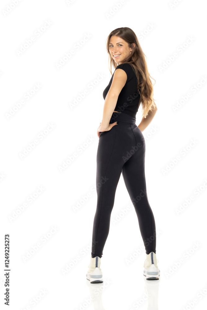 Smiling woman in black activewear posing sideways, hand on hip, wearing white sneakers on white background Smiling woman in black activewear posing sideways, hand on hip, wearing white sneakers on white background
