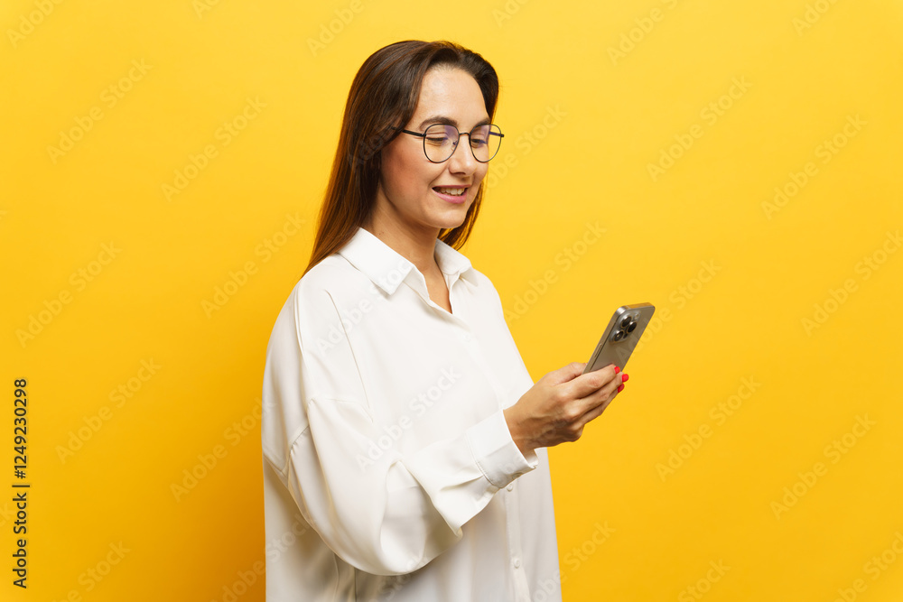Woman with glasses smiles while using smartphone against bright yellow background Woman with glasses smiles while using smartphone against bright yellow background