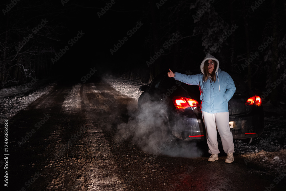 Obraz premium Woman stands next to her broken-down car on a snowy forest road at night, signaling for help. The cold winter scene is illuminated by the car's rear lights and rising steam.