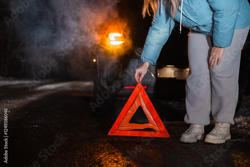 On a dark winter road, a woman places a reflective warning triangle beside her broken-down car. The scene highlights the importance of safety precautions during nighttime travel.