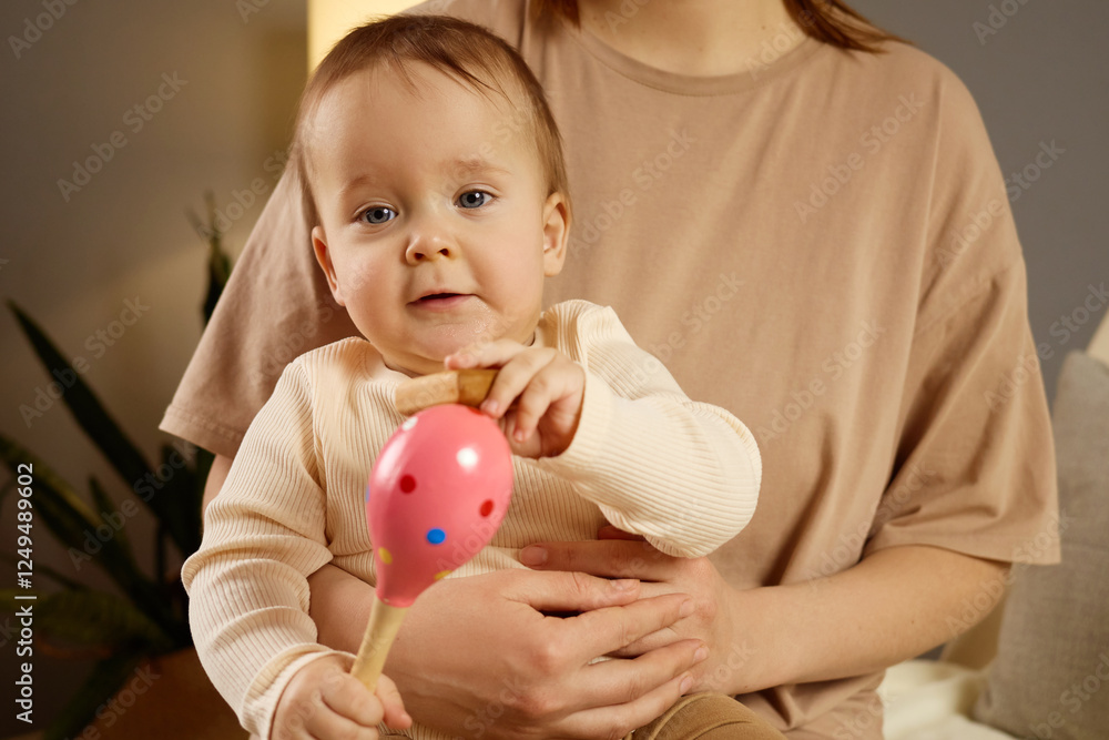 A baby with light brown hair is happily holding a pink wooden toy shaped like a balloon. Seated on a caregiver's lap, they are surrounded by a warm and inviting environment A baby with light brown hair is happily holding a pink wooden toy shaped like a balloon. Seated on a caregiver's lap, they are surrounded by a warm and inviting environment