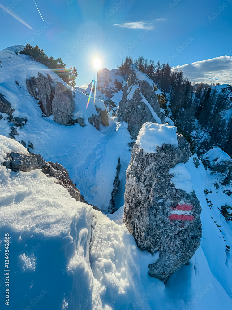 Sunlight illuminates snow-covered rocks in Mallestiger Mittagskogel, Austria. Alpine trail mark guides hikers through extreme winter landscape. Bright, clear day in alpine wildereness of Austrian Alps