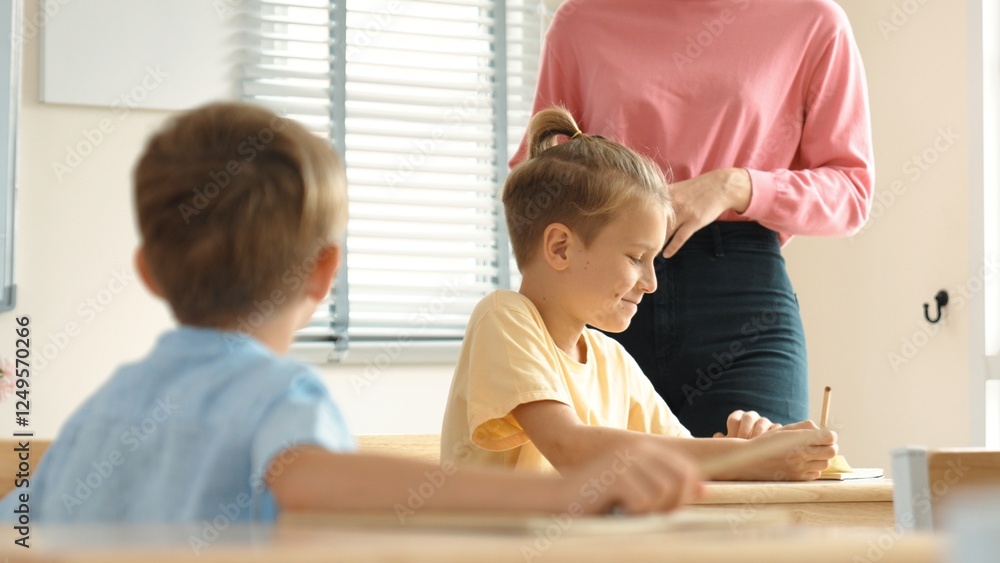 Fototapeta premium Beautiful teacher explaining test while caucasian boy doing classwork and listening explanation. Cute child looking at teacher while taking a notes or writing answer in paper at classroom. Pedagogy.