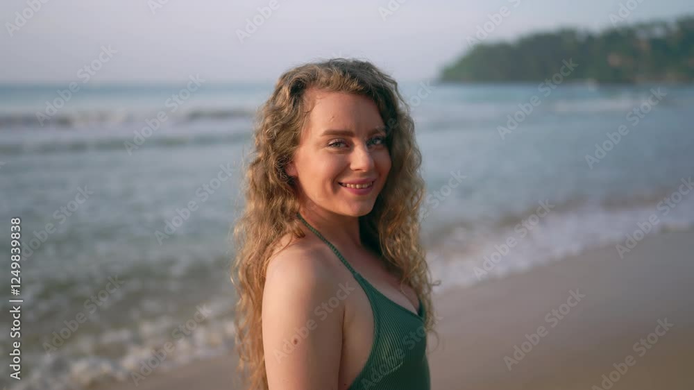 Curly-haired woman in green swimsuit walks along tropical beach, waves gently breaking on shore behind. Female enjoys serene seaside, displays lustrous hair, exemplifies travel leisure.