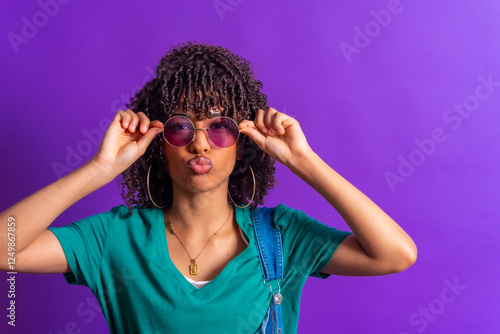Young woman adjusting sunglasses and pouting on purple background