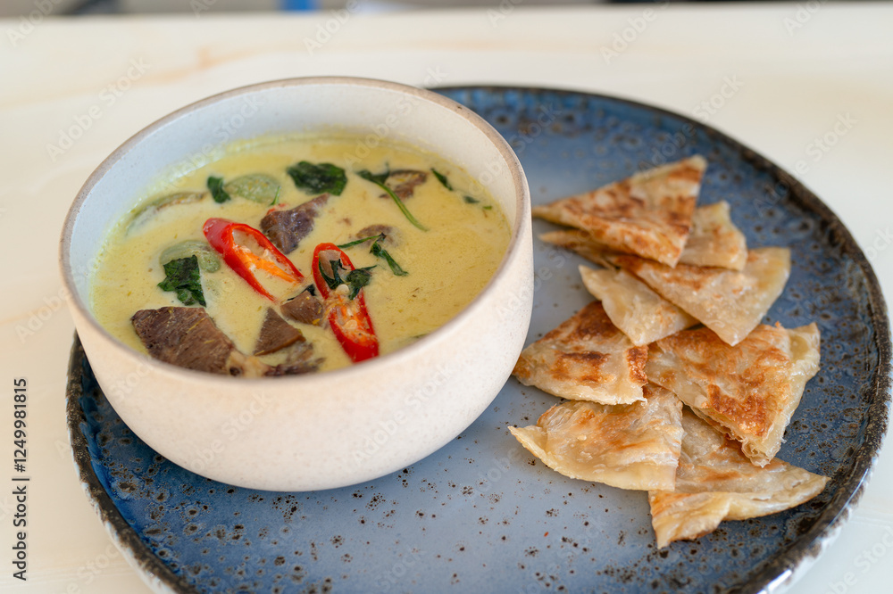 A serving of green curry in a white bowl, placed on a wide, flat dish with a portion of crispy roti canai. A serving of green curry in a white bowl, placed on a wide, flat dish with a portion of crispy roti canai.