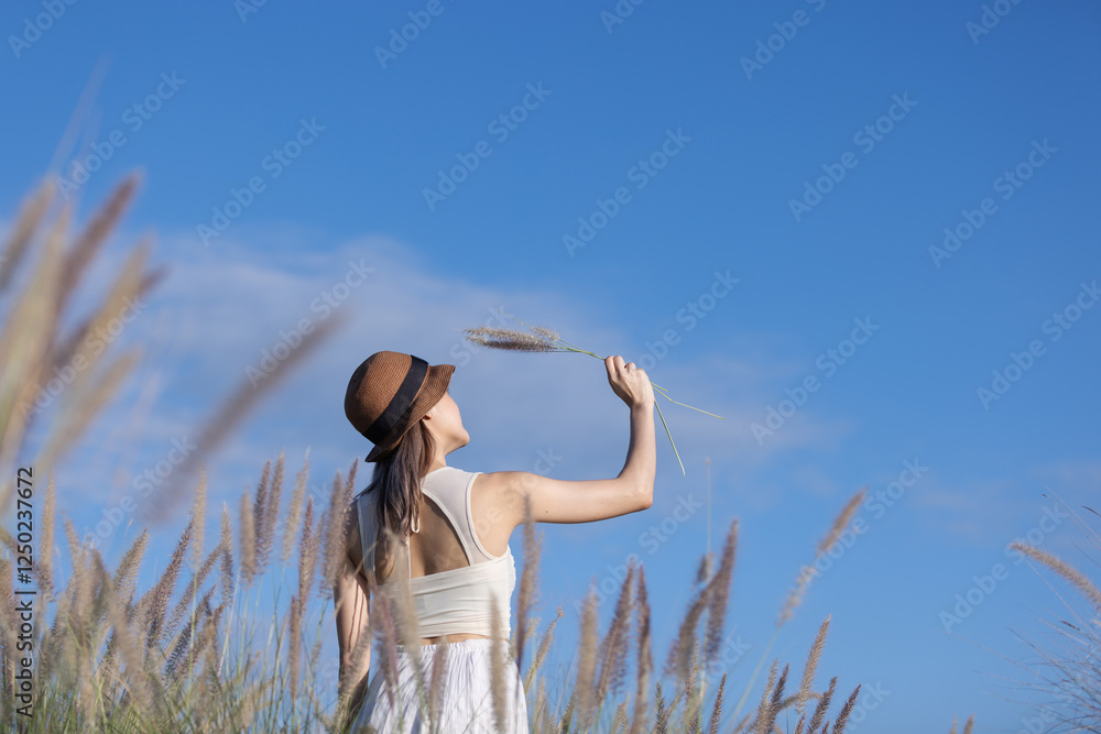 A young woman in a white dress and hat stands in a field, raising her arms toward the clear blue sky while holding stalks of grass. A young woman in a white dress and hat stands in a field, raising her arms toward the clear blue sky while holding stalks of grass.
