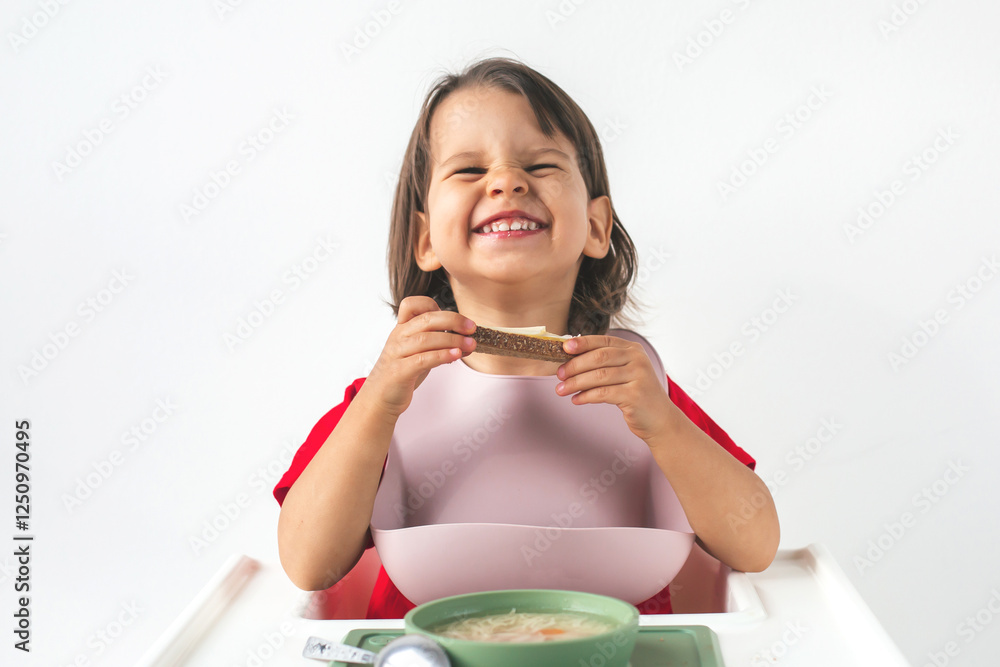 Cheerful little girl with dark hair sitting in a high chair, grinning widely while holding a slice of bread with cheese, silicone bib and enjoying her meal. Concept of childhood, healthy eating Cheerful little girl with dark hair sitting in a high chair, grinning widely while holding a slice of bread with cheese, silicone bib and enjoying her meal. Concept of childhood, healthy eating