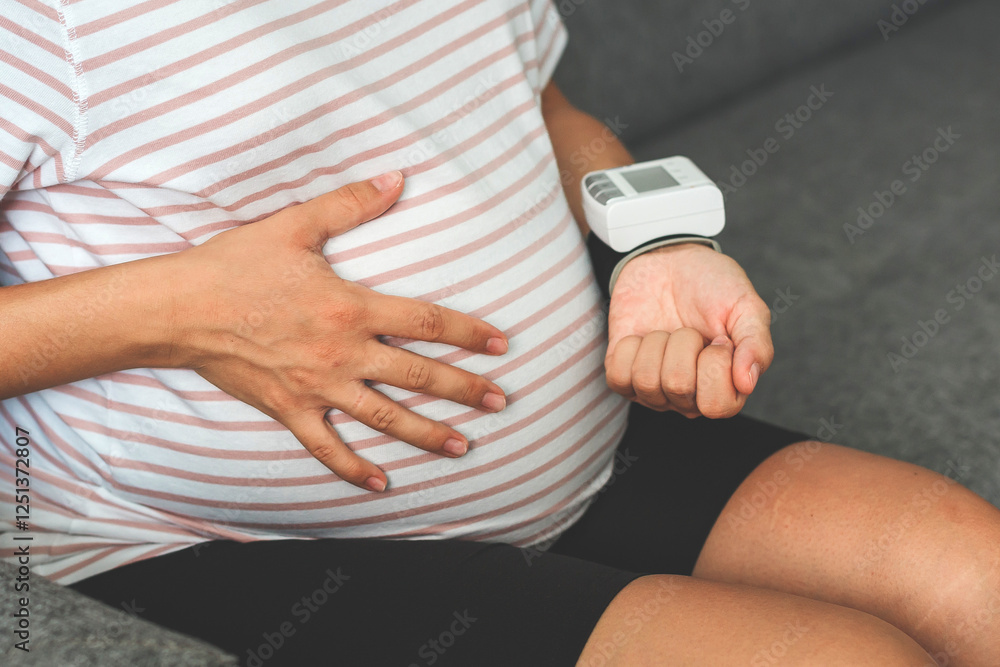 Obraz premium Pregnant woman in a striped t-shirt measuring blood pressure with a wrist monitor while sitting on a couch, holding her belly with one hand, focusing on health and well-being
