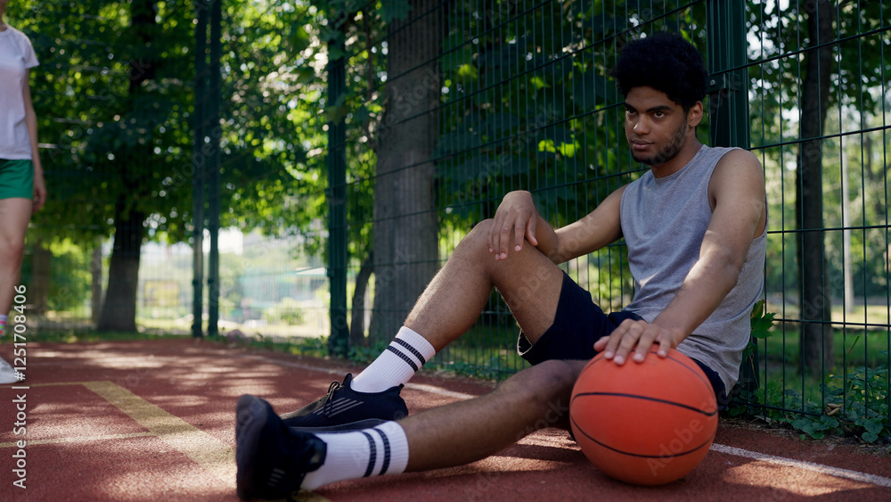 African American young man rests after playing basketball, waiting for his companions on the court African American young man rests after playing basketball, waiting for his companions on the court