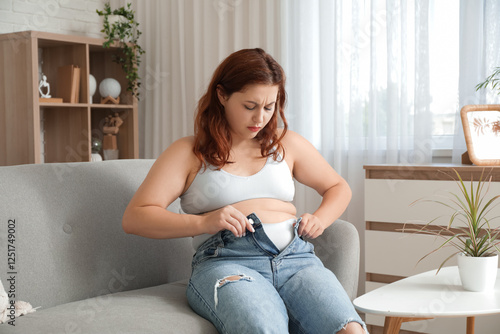Overweight young sad woman trying to button tight jeans at home. Weight gain ...