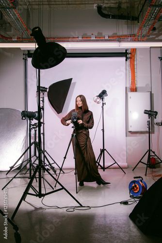 A female photographer at work in a photo studio with a professional camera on a tripod, lighting equipment on stands, a white backdrop, softbox, beauty dish, reflector. Stylish photo studio concept