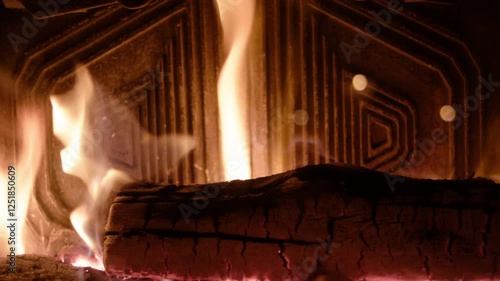 In the fireplace, flames sway against geometric metal panels with concentric shapes. A slowly charring log reveals glowing embers in its cracks, creating depth and mesmerizing warmth.